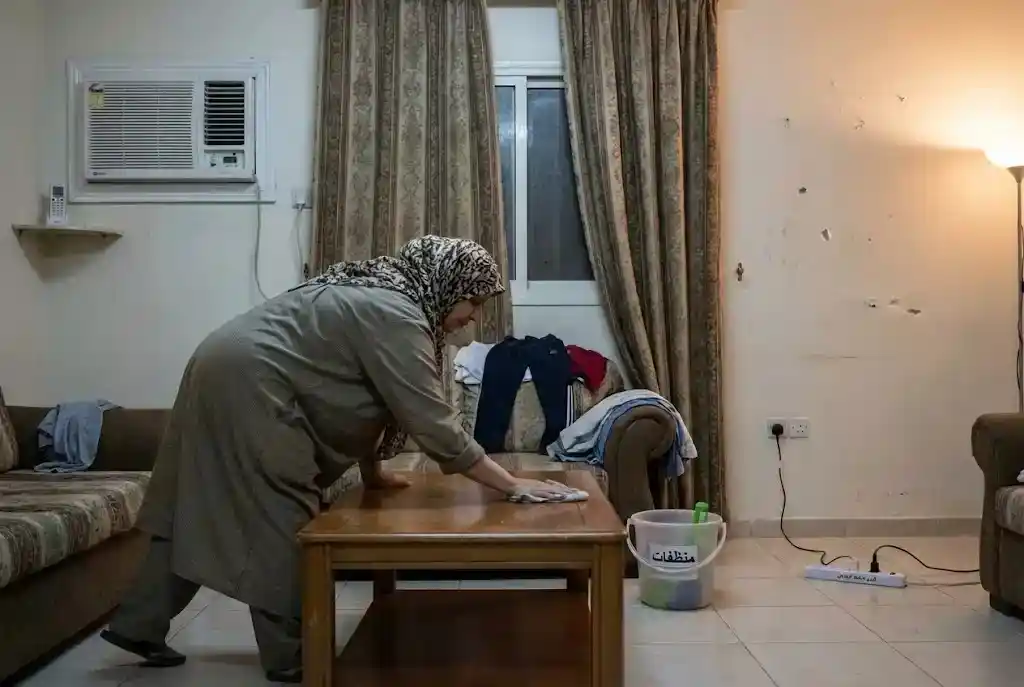 عاملة تنظيف تمسح طاولة في صالة بيت، مع كنبة وستائر ومكيف شباك، ودلو أدوات تنظيف على الأرض، في أجواء منزلية عادية